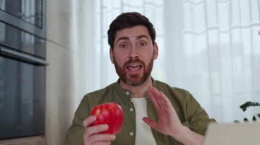 Beyaz tenli erkek vloger içeride kolları kıvrılmış yeşil gömlek giyen taze kırmızı elmanın besinlerinden bahsediyor. Beyaz perdeli ev işyeri ve pencerenin arka planı.