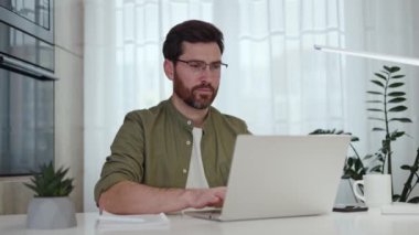 Modern oturma odasında dizüstü bilgisayarla oturan, başını çevirip kameraya gülümseyen yakışıklı sakallı adamın portresi. Mutlu erkek rahat iş yerinde rahat kıyafetler ve gözlük takıyor..