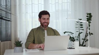 Laptop, içecek, lamba ve saksı bitkisiyle masada otururken rahat sandalyeye yaslanan sakallı adam. Kafkas işçilerinin zor işleri bitirmesi ve mutlu görünmeleri..