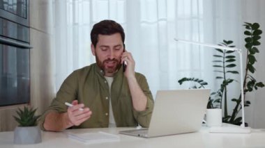 Yetişkin bir adam evde dizüstü bilgisayarla otururken telefonla konuşuyor ve gülümseyerek notlar alıyor. Beyaz erkek, iş yerinde meslektaşıyla yeni proje hakkında ayrıntıları tartışıyor.