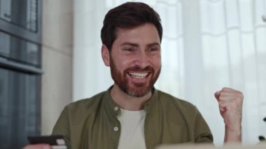 Gülümseyen erkek yönetici ev tabanlı iş ortamında kredi kartından kablosuz bilgisayardan para kontrol ediyor. Neşeli Kafkas çalışanı masada oturuyor ve memnuniyetten evet hareketi yapıyor..