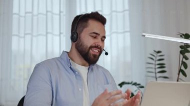 Kulaklıklı genç bir iş adamı dizüstü bilgisayarla video görüşmesi yapıyor ve ev içi müşterilerin sorularını cevaplıyor. Konsantre olmuş erkek danışman müşteri çevrimiçi ve satılık ürün sunar.