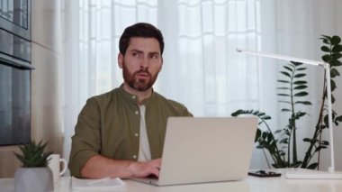 Profesyonel dizüstü bilgisayar kullanarak sakallı ve bıyıklı bir adam düşünceli bir şekilde yana bakıyor. Konsantre uzaklık çalışanı bir süre için şüpheye düştü ve proje başarısı için planlar geliştirmeye devam ediyor.
