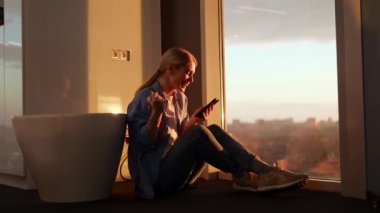 Atkuyruklu sarışın, duygusal bir kadın çatı katında oturmuş, elinde akıllı bir telefon tutarken çok heyecanlanıyor. Kadın aygıt kullanıcısı uzun zamandır bekletilen mesajı alıyor ve telefondan göğse tutunuyor.