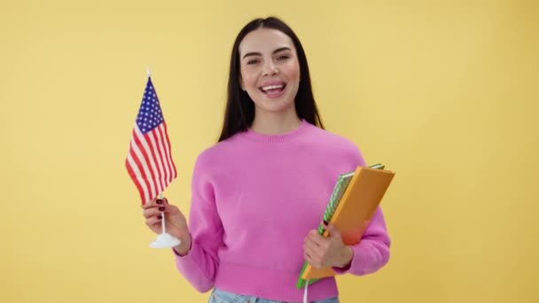 一个乐观的女人手里拿着书 一边挥动着美国国旗 一边站在有黄色背景的工作室里 穿着休闲装的快乐女生支持国庆并感到兴奋 — 图库视频影像