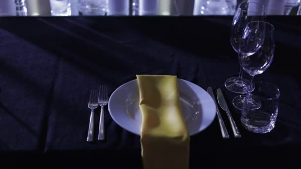 カトラリー付きのエレガントな空のテーブルセッティング 上部に黄色のナプキンとワインと水のメガネ付きの白いプレート 人工照明付きの豪華なレストランで1人のための黒いテーブルクロスでのサービス — ストック動画