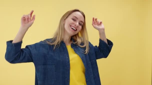 陽気で魅力的な女性の肖像画は 孤立した黄色の背景に対して手を上げて楽しんでいます スタジオセッティングで幸せを表現するデニムカジュアルジャケットを着た女性 — ストック動画