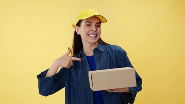 黄色い背景の小包箱に指を当てた誠実な笑顔の女性配達労働者 便利なライフスタイルのための配達サービスを促進する帽子とデニムシャツを身に着けている白人女性 — ストック動画