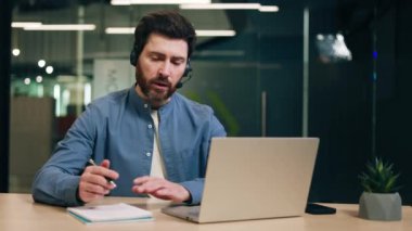 Kablosuz kulaklık takan, konuşurken el kol hareketi yapan ve dizüstü bilgisayar ekranına bakan meşgul bir adam. Günlük gömlekli Freelancer online video görüşmesi sırasında iş hakkında konuşuyor..