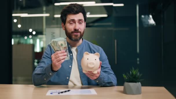 書類付きの机で座っている間 現金と古典的なマネーボックスのパックを握っている自信のある笑顔のブローカー 笑顔の男は 個人的な将来の資金を構築するためにクライアントに契約に署名することを助言 — ストック動画