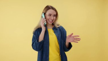 Duygusal kadın, izole edilmiş sarı arka planda dikilirken kendi telefonuyla enerjik bir konuşma yapıyor. Stüdyoda iletişimi ve modern teknolojileri seven tatmin olmuş bir kadın..