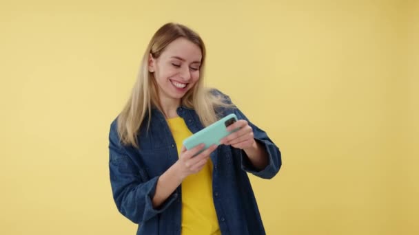 モダンモバイルでオンラインビデオゲームを熱狂的にプレイする感情的な面白い女性 黄色いスタジオの背景に隔離されたドライブシミュレータを使用してスマートフォンアプリを使用して幸せな陽気なかなり女性 — ストック動画