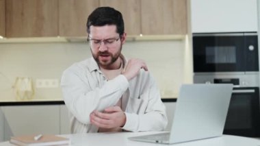Stresli bir işçi, kas gevşemesi için dirsek dirsek ve kemik ağrısı çekiyor. Sakallı adam, mutfak alanında modern taşınabilir bilgisayar kullanırken şiddetli acıdan gözlerini kısarak bakıyor..