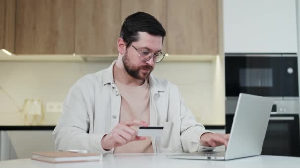 快乐的高加索男性手握信用卡在手提电脑上上网购物 坐在厨房内部的现代小玩意前 戴着眼镜的快乐大胡子的男人付了钱 — 图库视频影像