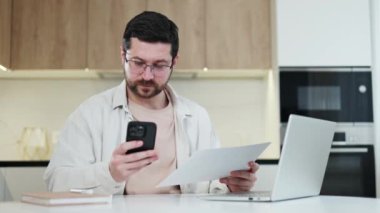 Kablosuz akıllı telefondan alınan belgelerle orijinalini karşılaştıran düşünceli beyaz erkek serbest çalışan. Düşünceli bir adam ev ortamındaki işiyle uğraşıyor. Modern mutfağın arka planı.