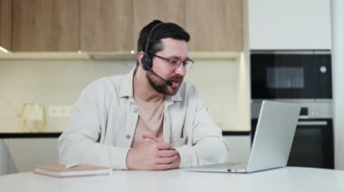 Tasarımcı mutfağının arka planında kendine güvenen bir adamın kişisel laptopuyla çevrimiçi konuşması. Kablosuz kulaklıklı gülümseyen beyaz adam interlocutorlarla konuşuyor..