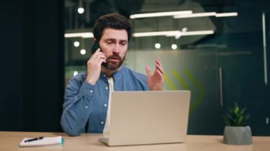 Sakallı, telefonla konuşan ve elleriyle hareket eden odaklanmış bir adam. Erkek destek ajanı müşterilere danışmanlık yapıyor ve iş yerinde dizüstü bilgisayarla otururken internet ile ilgili teknik sorunların çözümüne yardımcı oluyor.