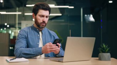 Ciddi bir erkek müdürün masamda dizüstü bilgisayarla oturup iş için akıllı telefon kullandığı bir portre. Koyu renk saçlı bir adam modern aletler tutuyor ve birlikte çalıştığımız yerde e-posta mektupları okuyor..