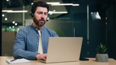 Dizüstü bilgisayarı olan ve şık bir ofiste çalışan sakin bir erkek çalışan. Kablosuz kulaklıkta profesyonel yönetici bilgisayar yazarken çağrı ve istemcinin sorunu çözmesine yardım ediyor.