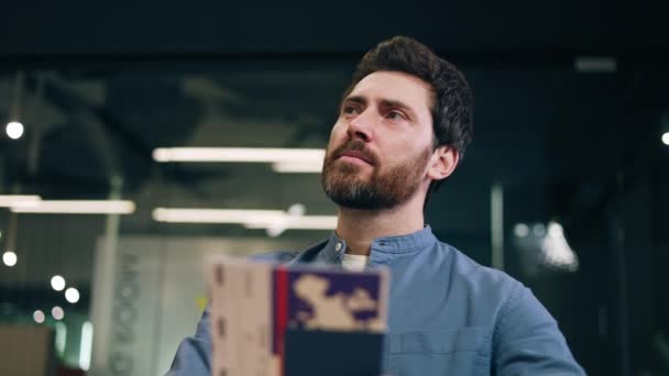 受到启发的男性穿着蓝色衬衫阅读小册子 带着沉思的微笑看着旁边模糊的背景 办公室的工作人员粗心大意地想象着在繁忙的一天里出去走走的情景 — 图库视频影像