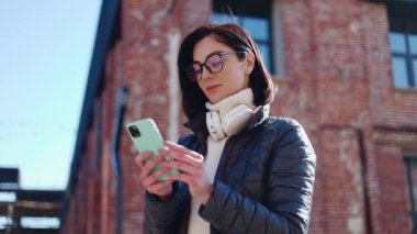 Açık mavi kutuda açık havada dururken modern akıllı telefonu kullanan koyu renk saçlı kadının portresi. Elektronik aygıta konsantre kadın metin yazma ve halka açık internet bağlantısı.
