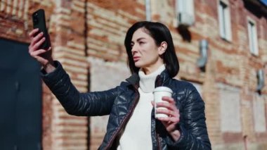 Milenyum kadını elinde plastik kahve fincanıyla yüksek açılı selfie çekerken mobil cihaz kullanıyor. Popüler yaşam tarzı etkileyicisi dışarıda yürürken sosyal medya için içerik üretiyor.
