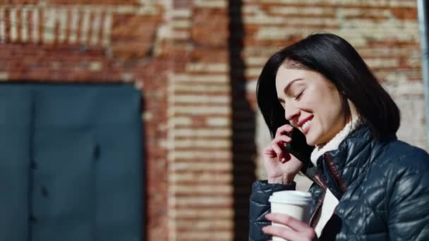 Urocza Kobieta Rozmawiająca Przez Smartfona Przechodząc Obok Ceglanego Budynku Kawą — Wideo stockowe