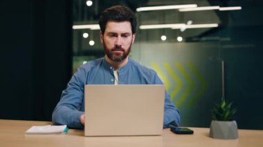 Laptopun önünde oturan ve rahat bir ofiste çalışan konsantre beyaz bir adam. Sorumlu erkek çalışan e-posta yazıyor ve masa olarak otururken kameraya bakmak için mola veriyor..