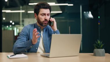 Modern ofis odasında portatif bilgisayarla otururken akıllı telefondan konuşan meşgul bir adam. Erkek girişimci ortaklarla iş görüşmesi yapıyor ve sözleşme detaylarını tartışıyor.