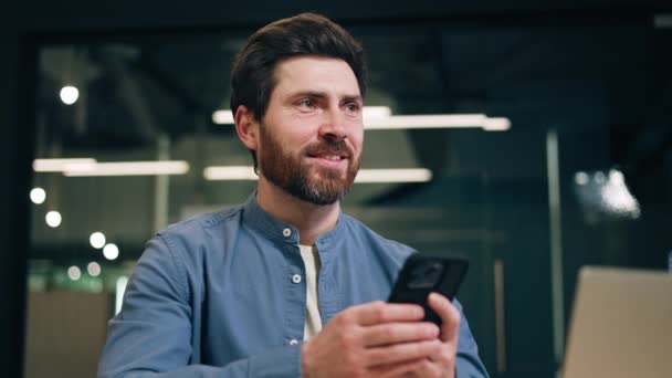 コワーキングスペースに座っている間 現代のスマートフォンと笑顔を使用してひげを持つ大人の男の近く 満足している男性マネージャーは インターネットを閲覧し プロジェクトについて考えながら素晴らしいアイデアを得る — ストック動画