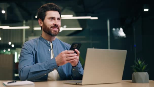 穿着时髦衬衫的男人在智能手机上打字 坐在电脑前笑着 男性员工在休息时使用现代设备进行娱乐和与朋友聊天 — 图库视频影像
