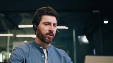 Kendine güveni tam bir erkek kulaklığı kontrol ederken dizüstü bilgisayarın önünde oturuyor. Dost canlısı destek ajanı müşterilere danışmanlık yapıyor ve modern ofiste önemli tavsiyelerde bulunuyor..