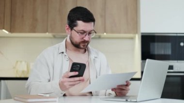 Belgeden modern akıllı telefona, kişisel çalışma alanı boyunca evde serbest çalışmak için belgeden modern akıllı telefona bakan yetkin beyaz bir erkek. Sakallı adam başarılı bir şekilde kapalı alandaki görevleri yerine getiriyor..