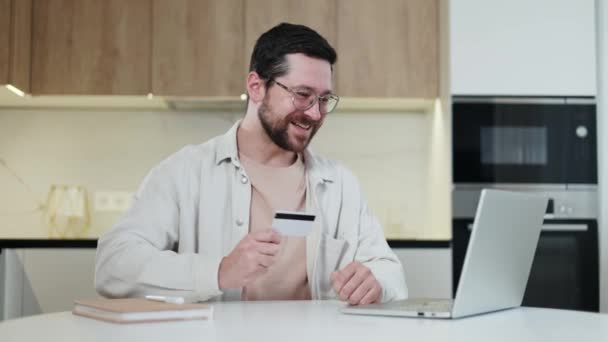快乐的男性持有塑料卡 在自己的银行户头里享受丰厚的利润带来的快乐 满头胡须的男子坐在打开的无线膝上型计算机前 目光炯炯地看着里面 — 图库视频影像