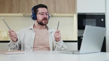 Masaüstünde bilgisayar ve defterle otururken kalem kullanarak mavi kulaklıklı beyaz bir erkek. Uzaktan çalışma günlerinde konserde rock yıldızı gibi davranan yetenekli bir adam..