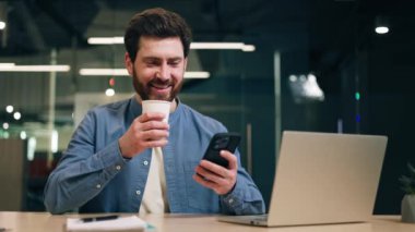 Erkek müdür kahve tadıyor ve gözlerini kapatıyor. Bu arada da çalışmaktan zevk alıyor. Ofis masasında oturup dizüstü bilgisayarda yazmaktan dinlenirken akıllı telefonu eğlence için kullanan rahat bir adam..