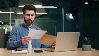 Erkek yönetici zarfı açıyor ve e-posta yoluyla olumlu haberler alırken gülümsüyor. Sakallı yakışıklı adam mektup okuyor ve modern dizüstü bilgisayarın ofisinde otururken memnuniyetini dile getiriyor..
