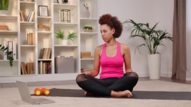 Genç Afrikalı Amerikalı kadın, modern dizüstü bilgisayarında online antrenörle yoga yapıyor ve hareketleri tekrarlıyor. Sakin kadın spor kıyafetleri giyiyor, rahat oturma odasında siyah yoga minderinin üzerinde rahatlıyor..