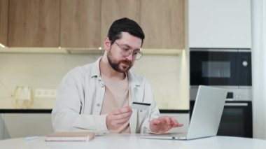 Sakallı, serbest çalışan, elinde kredi kartı tutan ve evdeki kablosuz bilgisayardan online alışveriş yapan biri. Gülümseyen beyaz bir erkek kendi parasıyla internet sitelerinden alışveriş yapıyor. Para ve insan kavramı.