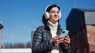 Yetişkin esmer kadın cep telefonuyla neşeli bir şarkı açıyor ve kulaklıkla ritimle dans ediyor. Dikkatsiz bayan dışarıda eğlenirken aynı zamanda favori müzik ve hareketlerle havasını düzeltiyor..