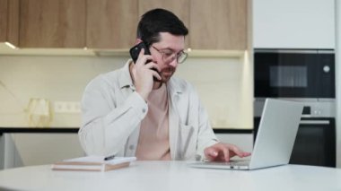 Kafkas erkek internet sitelerine kablosuz laptopla bakarken mutfak içinde kişisel akıllı telefonuyla konuşurken yan görünüm. Günlük kıyafetler ve gözlük takan gülümseyen adam iş yerinde vakit geçiriyor..