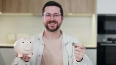 Gülümseyen esmer adam elinde domuz şeklinde para kutusu ve metal para tutarken kameraya tatmin olmuş bir yüz ifadesiyle bakıyor. Gelecek planlar için para biriktirme metodunu seçen kendinden emin bir adam..