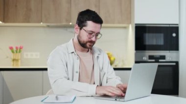 Modern laptop klavyesine tıklayıp doğrudan kameraya bakarken iş yerinde oturan yakışıklı bir gencin ön görüntüsü. Sakallı, beyaz, uzaktan kumandayla çalışan bir erkek..