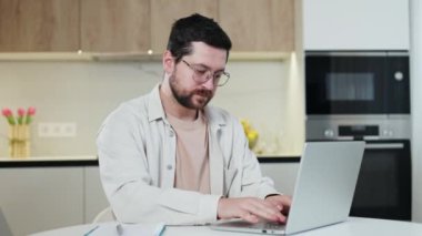 Düşünceli beyaz erkek serbest çalışanın ev ortamında kişisel dizüstü bilgisayarında uzak mesafe görevleri yapmasının yan görüntüsü. Yakışıklı, gözlüklü ve gündelik kıyafetleri olan bir adam modern aletler üzerinde yoğun bir şekilde çalışıyor..