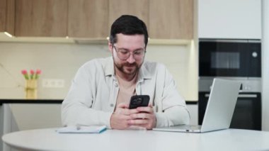 Bilgisayarlı ve defterli masada otururken, gözlüklü, modern akıllı telefona göz atan düşünceli beyaz bir erkek. İyi ışıklandırılmış ev ofisinde mesai saatinde dinlenmek için mola veren düşünceli kadın..