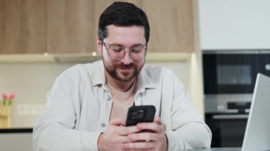 Ahşap mutfak dolaplarının arka planında dinlenirken kişisel akıllı telefonuyla sosyal medyada gülümseyen beyaz erkek görüntülerini kapatın. Düşünceli adam bağlantıyı koparmamak için modern aletler kullanıyor..