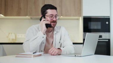 Olumlu Kafkasyalı serbest yazar evden uzaktan online iş yapıyor ve müşteriyle kapalı mekanda modern akıllı telefon aracılığıyla konuşuyor. Gülümseyen yakışıklı erkek ev ortamında iş sorunları çözüyor.