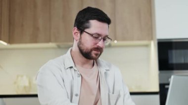 Neşeli beyaz erkek kişisel dizüstü bilgisayarında yazı yazıyor ve uzaktan çalışırken kameraya bakıyor. Mutfakta günlük giysiler ve gözlük takan pozitif sakallı bir serbest çalışan..