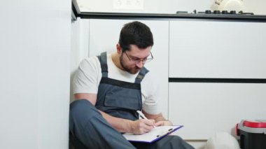 Mutfak zemininde oturan ve boyunu santimetre bantla ölçen gözlüklü profesyonel bir işçi. Yoğunlaştırılmış tamirci, panoya yenisini koymak için drenaj sisteminin göstergelerini yazıyor.