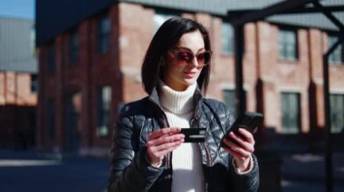 Sıradan kıyafetli olumlu bir kadın, plastik kart bilgilerini şehir arka planında dokunmatik ekranlı akıllı telefona kopyalıyor. Çekici bir kadın çevrimiçi alışveriş seansını tamamlıyor ve nakit ödemeyi onaylıyor..